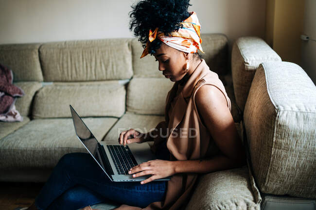 Высокий угол бокового обзора афроамериканки в стильном наряде, сидящей дома на диване и сидящей на нетбуке во время работы над удаленным проектом — стоковое фото