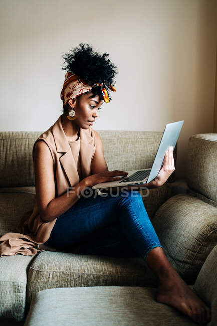 Side view of African American female in stylish outfit sitting on sofa at home and typing on netbook while working on remote project — Stock Photo