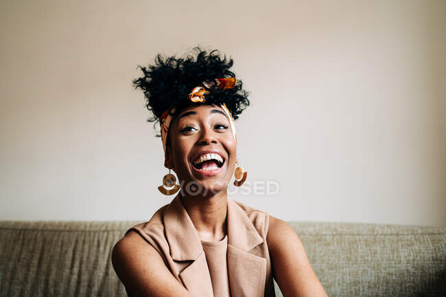 Веселая афроамериканка с модной повязкой на голове и прической в стиле афро, сидящая на диване и смеющаяся, глядя вдаль на дом — стоковое фото