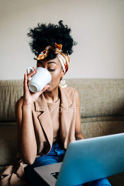 Ocupada mujer afroamericana freelancer sentada en el sofá con el ordenador portátil y beber café de la taza mientras trabaja en el proyecto en línea - foto de stock