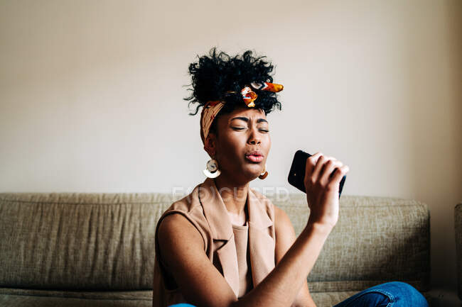 Carefree African American female sitting on sofa and singing favorite song in smartphone while enjoying weekend at home — Stock Photo