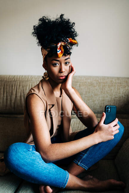 Fiduciosa donna afroamericana con fascia alla moda e con acconciatura afro seduta sul divano e appoggiata a portata di mano mentre guarda la fotocamera a casa — Foto stock