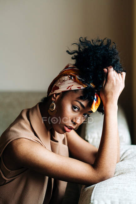 Mujer afroamericana segura con diadema de moda y con peinado afro sentado en el sofá y apoyado en la mano mientras mira la cámara en casa - foto de stock