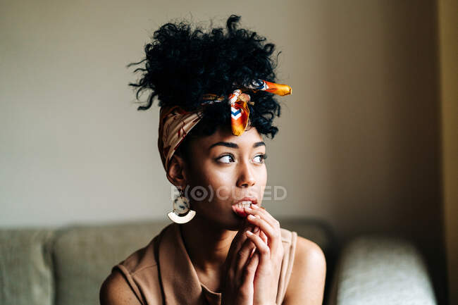 Fiduciosa donna afroamericana con fascia alla moda e con acconciatura afro seduta sul divano e appoggiata a portata di mano mentre guarda lontano a casa — Foto stock