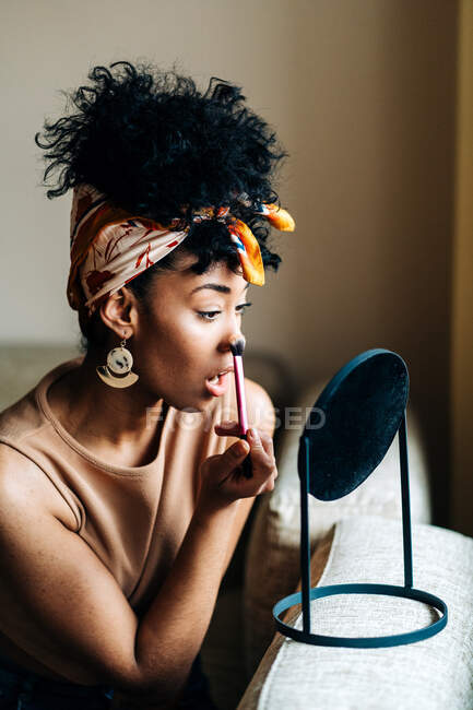 Vue latérale de charmante femme afro-américaine regardant dans un miroir rond et appliquant de la poudre sur le visage tout en faisant du maquillage à la maison — Photo de stock