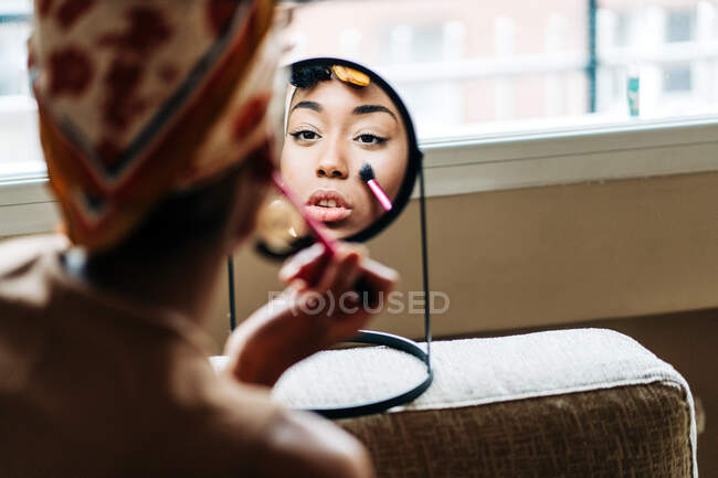 Vista posterior de la encantadora hembra afroamericana mirando en espejo redondo y aplicando polvo en la cara mientras hace maquillaje en casa - foto de stock