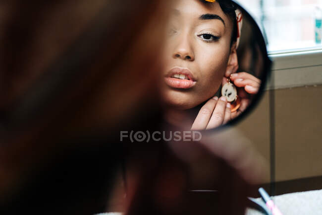 Reflection of content African American female putting on earring and looking in round mirror at home — Stock Photo
