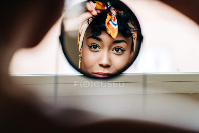 Reflexão do conteúdo afro-americano feminino colocando na cabeça e olhando no espelho redondo em casa — Fotografia de Stock