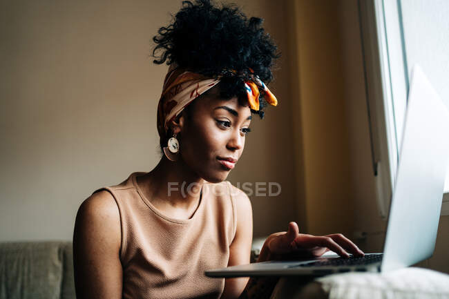 Афроамериканська жінка в стильному одязі сидить вдома на дивані і друкує на нетбуку під час роботи над віддаленим проектом. — стокове фото