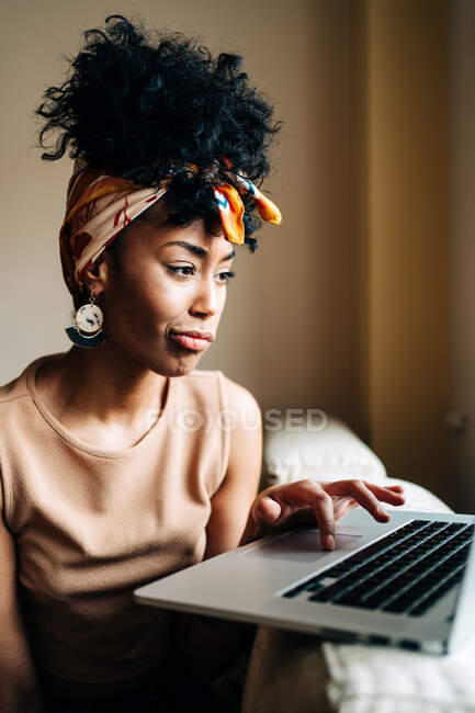 Mujer afroamericana en traje elegante sentada en el sofá en casa y escribiendo en netbook mientras trabajaba en un proyecto remoto - foto de stock