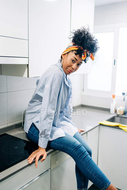 Vista lateral de la mujer afroamericana con el pelo rizado sentado en el mostrador en la cocina moderna y mirando a la cámara - foto de stock