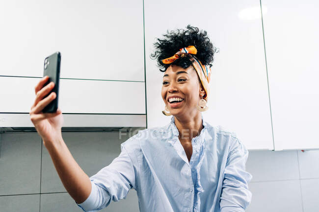 Веселая чёрная домохозяйка в повязке на голове делает автопортрет на смартфоне на кухне дома — стоковое фото