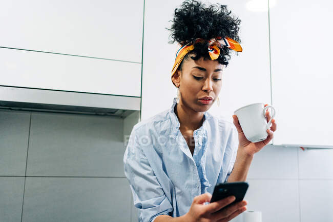 Afroamericana mujer navegando por Internet en el teléfono inteligente mientras bebe café en la cocina por la mañana - foto de stock
