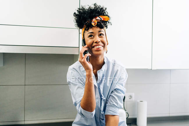 African American female speaking Internet on smartphone while siting in countertop in the kitchen in morning — Stock Photo