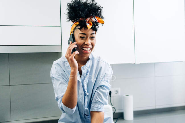 Mujer afroamericana hablando Internet en el teléfono inteligente mientras se encuentra en la encimera en la cocina por la mañana - foto de stock