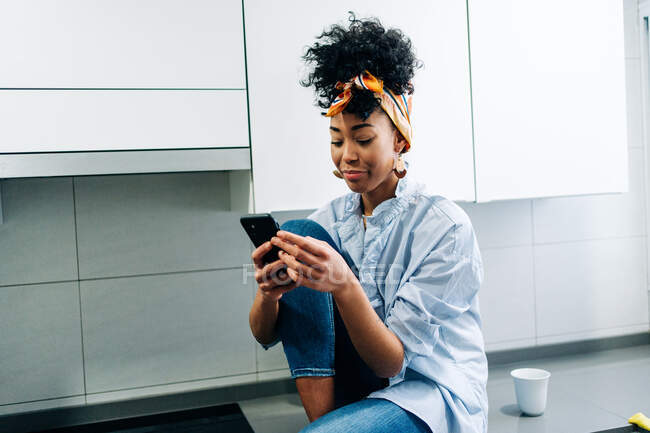 Afro-americano feminino navegar na Internet no smartphone, enquanto se situa na bancada na cozinha pela manhã — Fotografia de Stock