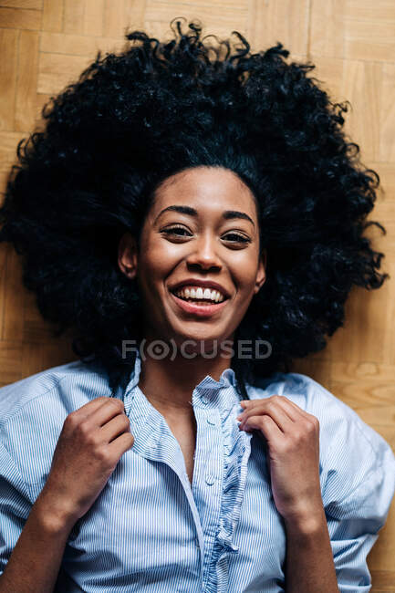 From above of slim African American female with curly hair lying on parquet floor and looking at camera — Stock Photo