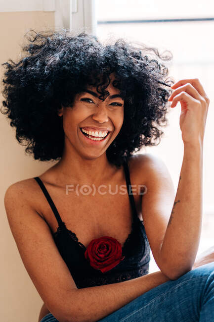 Optimistic African American female with rose flower in tank top touching curly hair and laughing at camera — Stock Photo