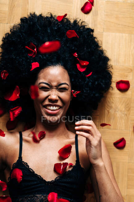 Draufsicht auf eine romantische, fröhliche Afroamerikanerin, die mit roten Rosenblättern auf dem Boden liegt, mit geschlossenen Augen — Stockfoto