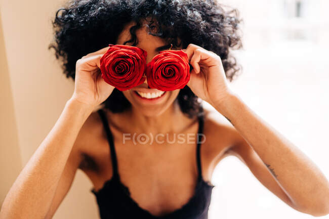 Alto ángulo de contenido Mujer afroamericana con peinado afro que cubre los ojos con brotes de flores de rosas rojas en casa - foto de stock