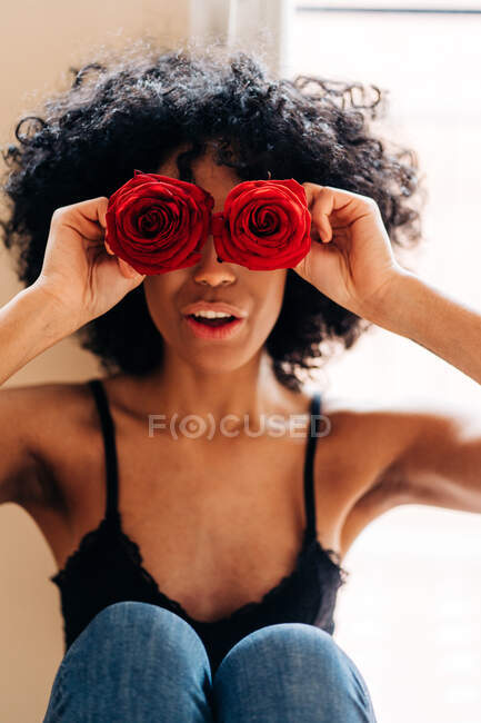High angle of content African American female with Afro hairstyle covering eyes with buds of red rose flowers at home — Stock Photo