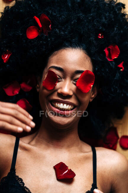 Vista superior de romántica mujer afroamericana alegre tumbada en el suelo con pétalos de rosa rojos con los ojos cerrados - foto de stock