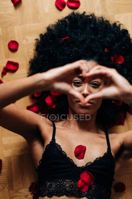 Draufsicht einer romantischen Afroamerikanerin, die mit roten Rosenblättern auf dem Boden liegt und Liebeszeichen zeigt, während sie in die Kamera schaut — Stockfoto
