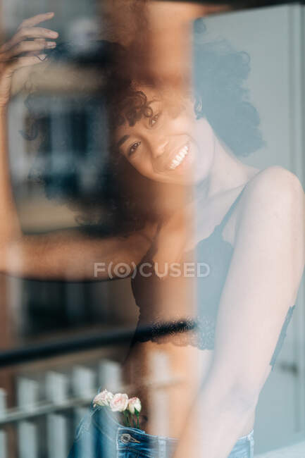 Par la fenêtre de femme afro-américaine ravie avec les cheveux bouclés debout avec des fleurs roses en jeans et en regardant la caméra — Photo de stock