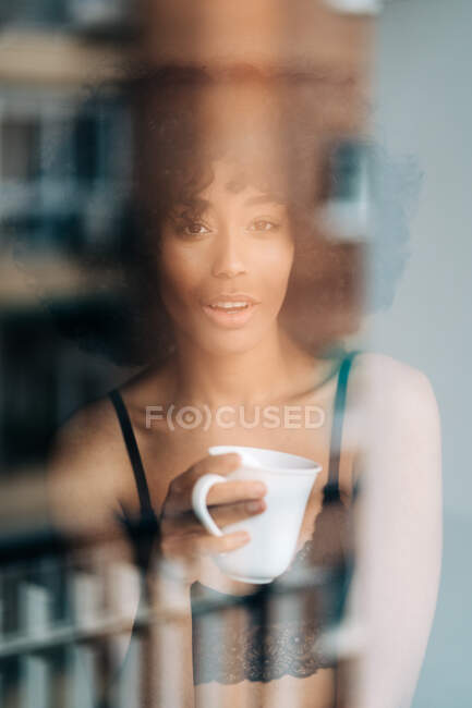 Através da janela de conteúdo afro-americano feminino em renda superior e com xícara de bebida olhando para a câmera — Fotografia de Stock