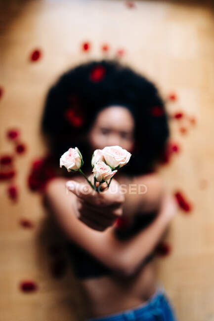 Von oben eine anonyme Afroamerikanerin, die mit verstreuten Blütenblättern auf dem Boden liegt und zarte Rosenblüten in die Kamera zeigt — Stockfoto