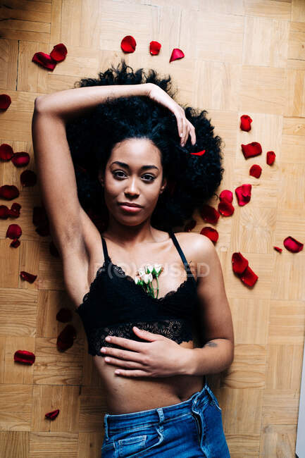 Von oben liegt eine schöne Afroamerikanerin auf dem Boden mit verstreuten Blütenblättern mit zarten Rosenblüten und blickt in die Kamera — Stockfoto