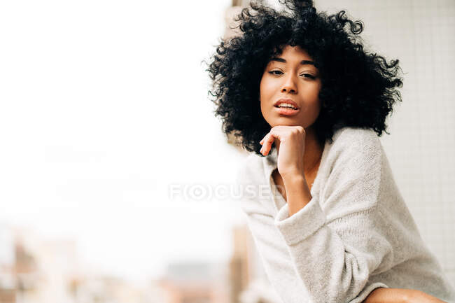 Убік погляд мирної афроамериканської жінки з кучерявим волоссям, що спирається на поручні, стоячи на балконі і дивлячись на камеру — стокове фото