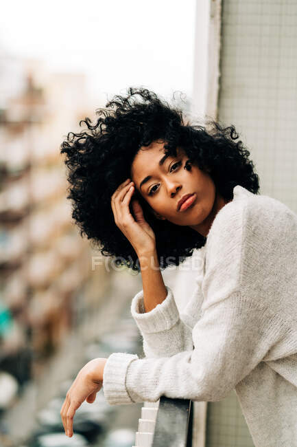 Vista laterale di pacifica donna afroamericana con i capelli ricci appoggiati sulla ringhiera mentre in piedi sul balcone e guardando la fotocamera — Foto stock
