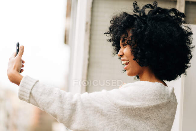Side view of charismatic black female with Afro hair standing on balcony and taking self shot on smartphone — Stock Photo