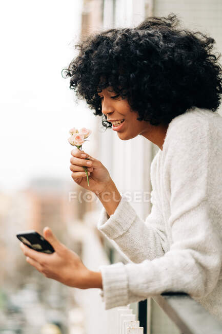 Vue latérale de la femme afro-américaine debout sur le balcon et prenant l'autoportrait avec des fleurs roses tout en utilisant un smartphone — Photo de stock