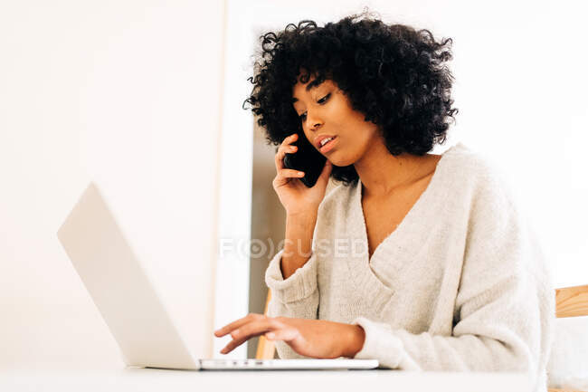 Seitenansicht einer viel beschäftigten afroamerikanischen Freelancerin, die am Tisch sitzt und an einem Projekt arbeitet, während sie am Laptop tippt und auf dem Handy spricht — Stockfoto