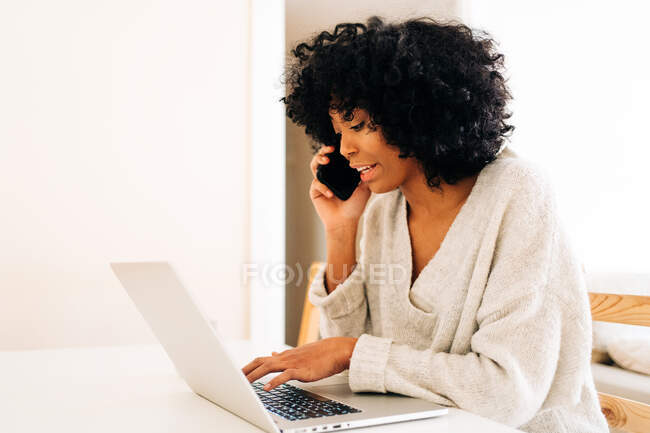 Vista lateral de la ocupada freelancer afroamericana sentada en la mesa y trabajando en el proyecto mientras escribe en el portátil y habla por teléfono móvil - foto de stock