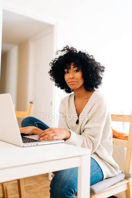 Délicieuse pigiste afro-américaine assise à table avec ordinateur portable et regardant la caméra pendant le travail à distance dans le bureau à domicile — Photo de stock