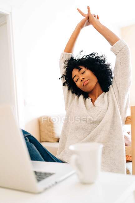 Уставшая афроамериканская предпринимательница, растягивающая руки, сидя за столом с ноутбуком и обедая дома на удаленной работе — стоковое фото