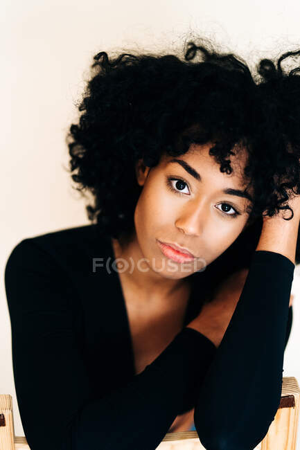 Sereno preto fêmea com cabelo encaracolado sentado na cadeira de madeira contra a parede branca e olhando para a câmera — Fotografia de Stock