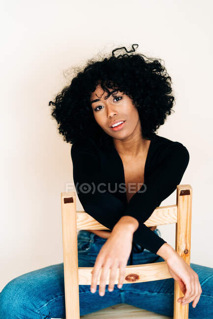 Черная женщина с кудрявыми волосами, сидящая на деревянном стуле у белой стены и смотрящая в камеру — стоковое фото