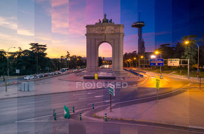 Arco de piedra con esculturas entre árboles y postes de luz iluminando carreteras con tráfico de transporte en Madrid al atardecer - foto de stock