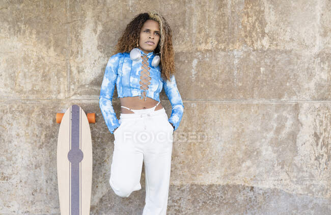 Trendy fiducioso giovane donna afroamericana in elegante camicia blu e pantaloni bianchi con le cuffie sul collo in piedi con le mani in tasche vicino skateboard contro muro di cemento shabby — Foto stock