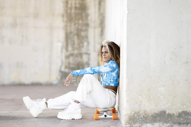 Vista lateral de cuerpo completo de la elegante joven hipster afroamericana en ropa de moda y botas sentadas en monopatín contra la pared de hormigón mientras descansa en la calle urbana - foto de stock