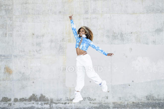 Pieno corpo di giovane donna hipster afroamericana ottimista in elegante abbigliamento informale e scarpe da ginnastica che saltano in alto sopra terra contro il muro di cemento squallido — Foto stock
