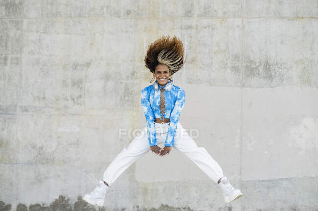 Corps complet de jeune femme afro-américaine hipster optimiste en tenue informelle élégante et baskets sautant haut au-dessus du sol contre un mur en béton minable — Photo de stock