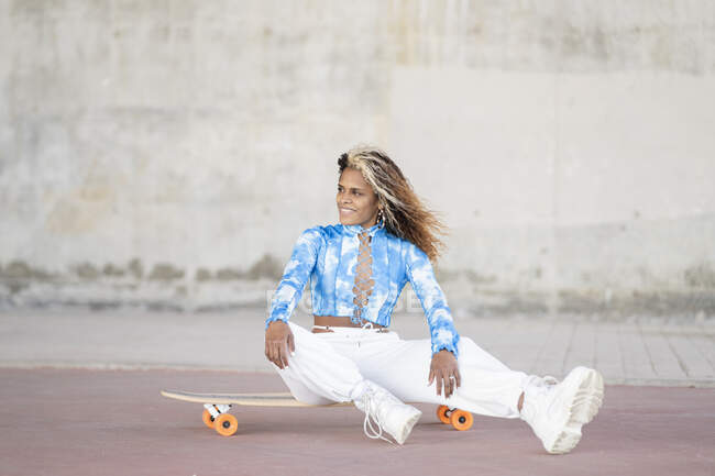 Все тело стильная молодая хипстерша афроамериканка в модной одежде и сапогах сидит на скейтборде у бетонной стены, отдыхая на городской улице — стоковое фото