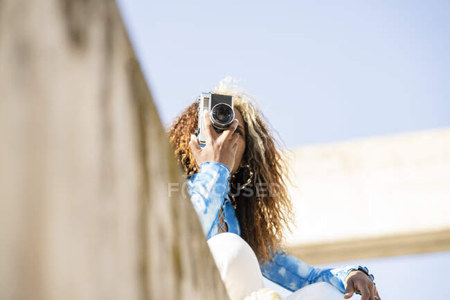Снизу счастливая кудрявая этническая женщина в стильном наряде сидит на бетонном заборе и фотографирует ретро-фотоаппаратом в солнечный летний день — стоковое фото
