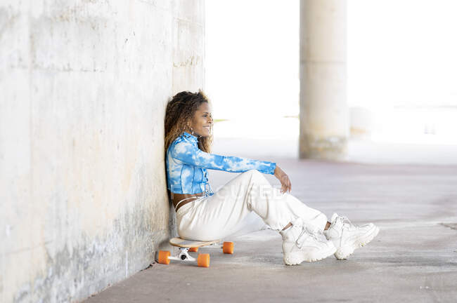 Vista lateral de cuerpo completo de la elegante joven hipster afroamericana en ropa de moda y botas sentadas en monopatín contra la pared de hormigón mientras descansa en la calle urbana - foto de stock