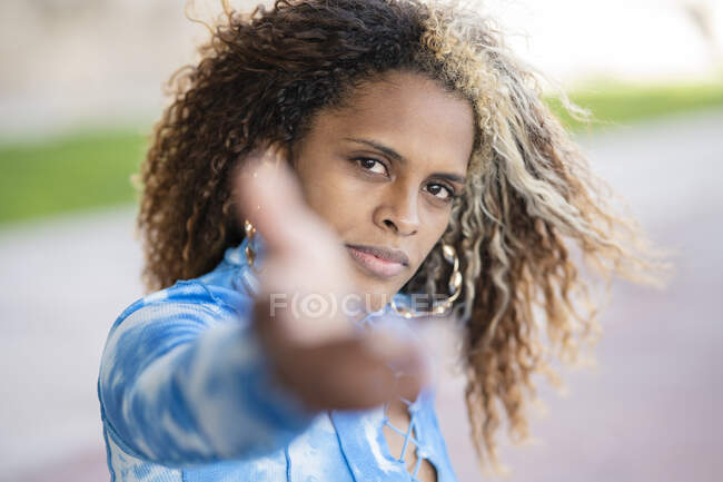 Впевнена сучасна афро-американська гіпстерка з кучерявою зачіскою і кільцевими сережками, одягнена в модний синій сорочці, розтягує руку і дивиться на камеру, стоячи на розмитій міській вулиці. — стокове фото